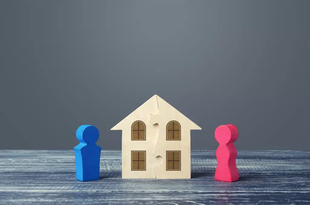 Zweiteiliges Holzhaus als Puzzle, getrennt durch eine Mittellinie, mit einer blauen und einer pinken Figur auf jeder Seite - symbolisch für die Aufteilung einer Immobilie beim Hausverkauf und die damit verbundenen steuerlichen Fragen.